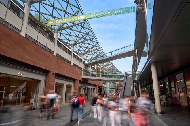 "Liverpool ONE walkway with shoppers, escalator to upper level and glazed roof structure overhead."