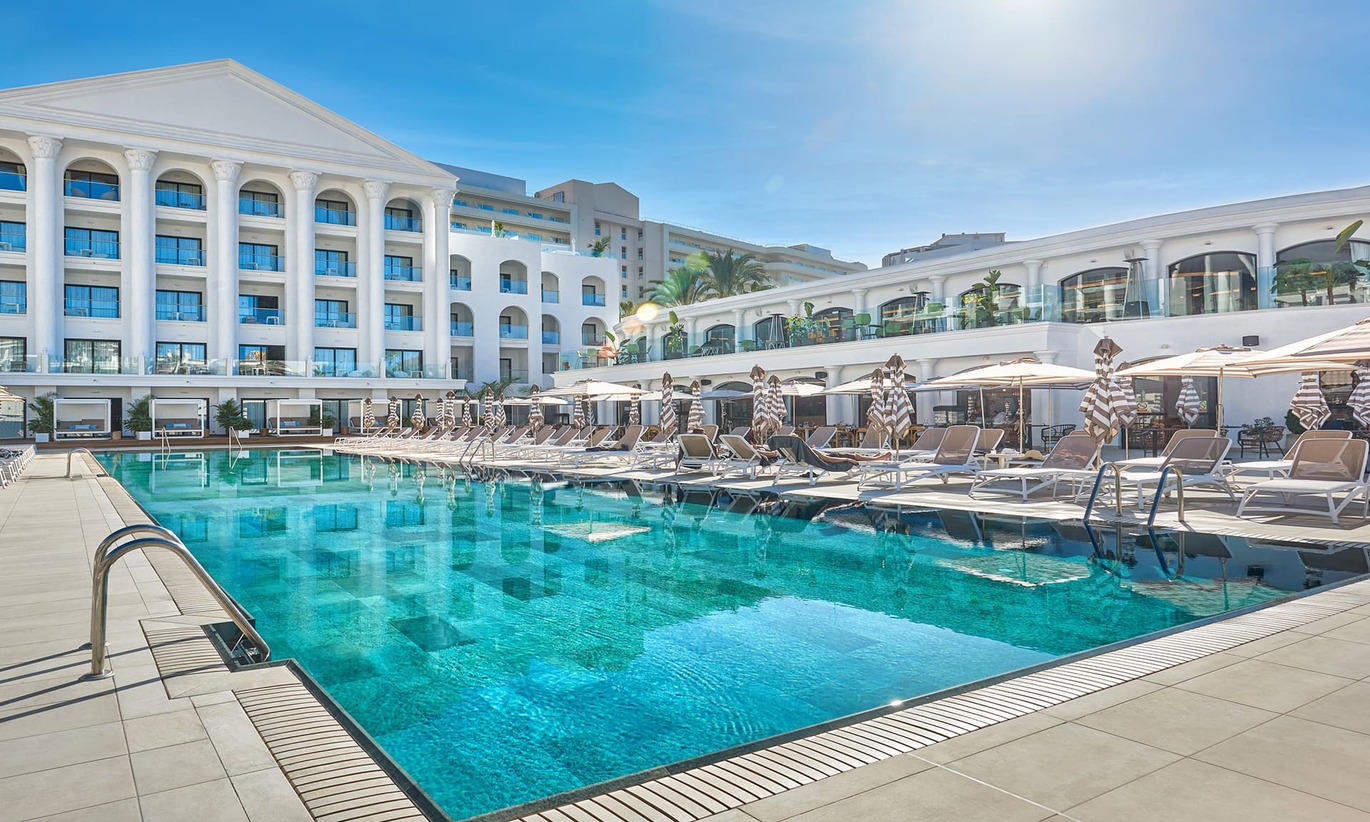 View from the back corner of the lovely clear pool with many sun loungers and umbrellas around the pool with the white hotel building surrounding the pool area on a lovely clear sunny day.