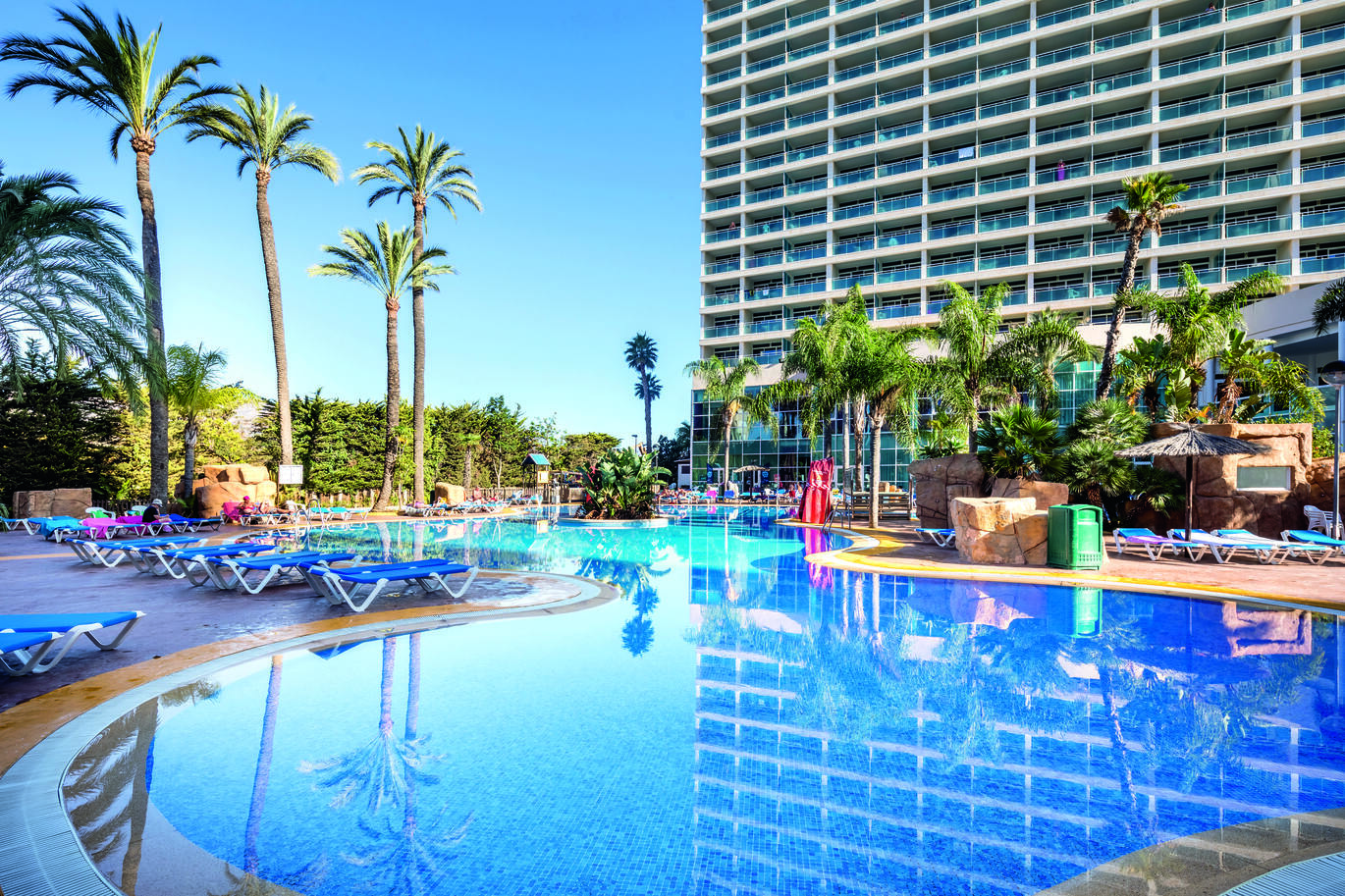 Hotel building in the background with palm trees and greenery in front with the pool in the centre with blue sun loungers around it.