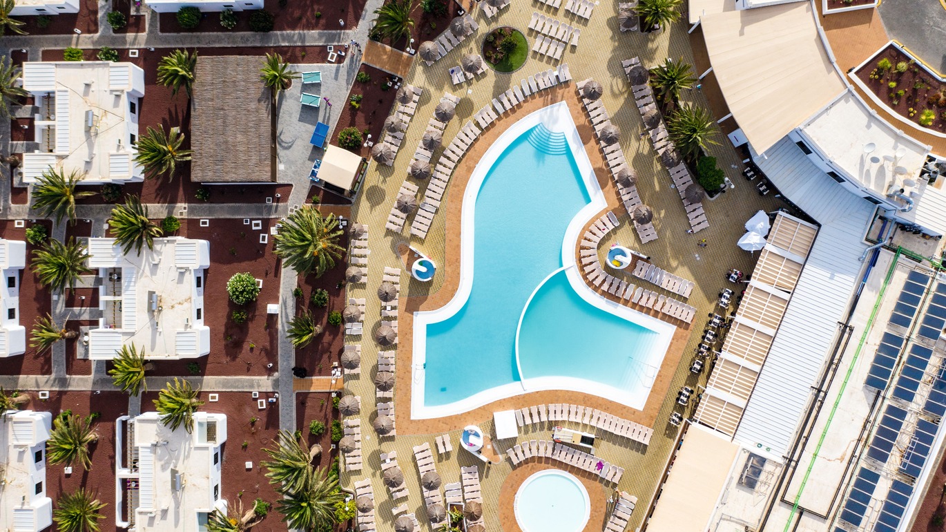 Birdseye view of part of the hotel complex with with the pool surrounded by many sun loungers and straw umbrellas.