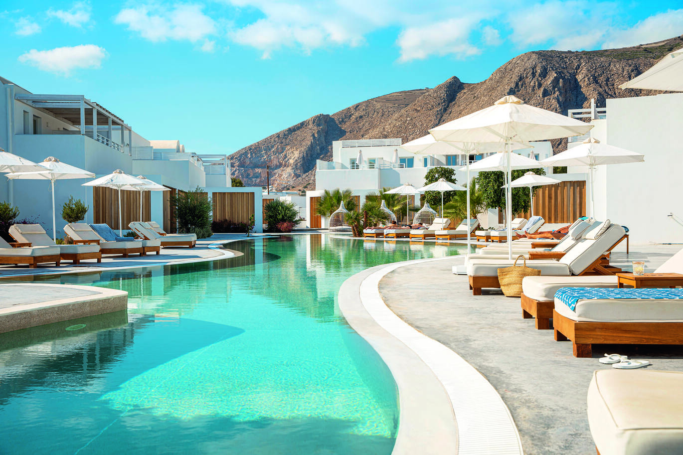 View of the pool area surrounded by sun beds and umbrellas with villa rooms behind and a view of the mountains in the background with the blue, slightly cloudy sky above.