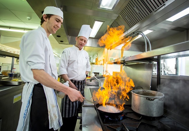 "Photo of 2 people cooking in a kitchen wearing chef uniforms. Chef is flambeeing food in pan"