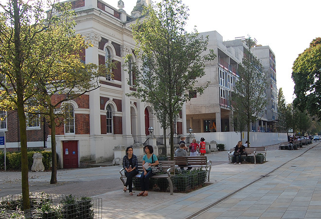 An open paved walkway with university buildings around the perimeter, two students are sat on benches next to some small trees