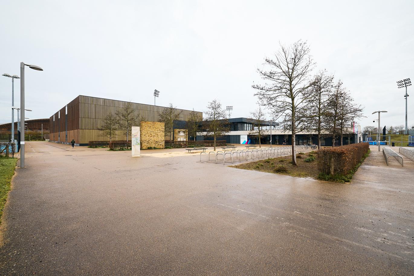 "Modern sports and hockey centre with timber-clad buildings, bike racks, and open paved forecourt with trees and pathways."