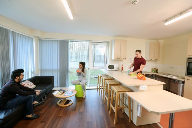"Kitchen in Talybont Court. A a height adjustable worktop is to the right foreground, with cooking facilities to the right rear. A student is cooking, with two other students sat down on the left in conversation"