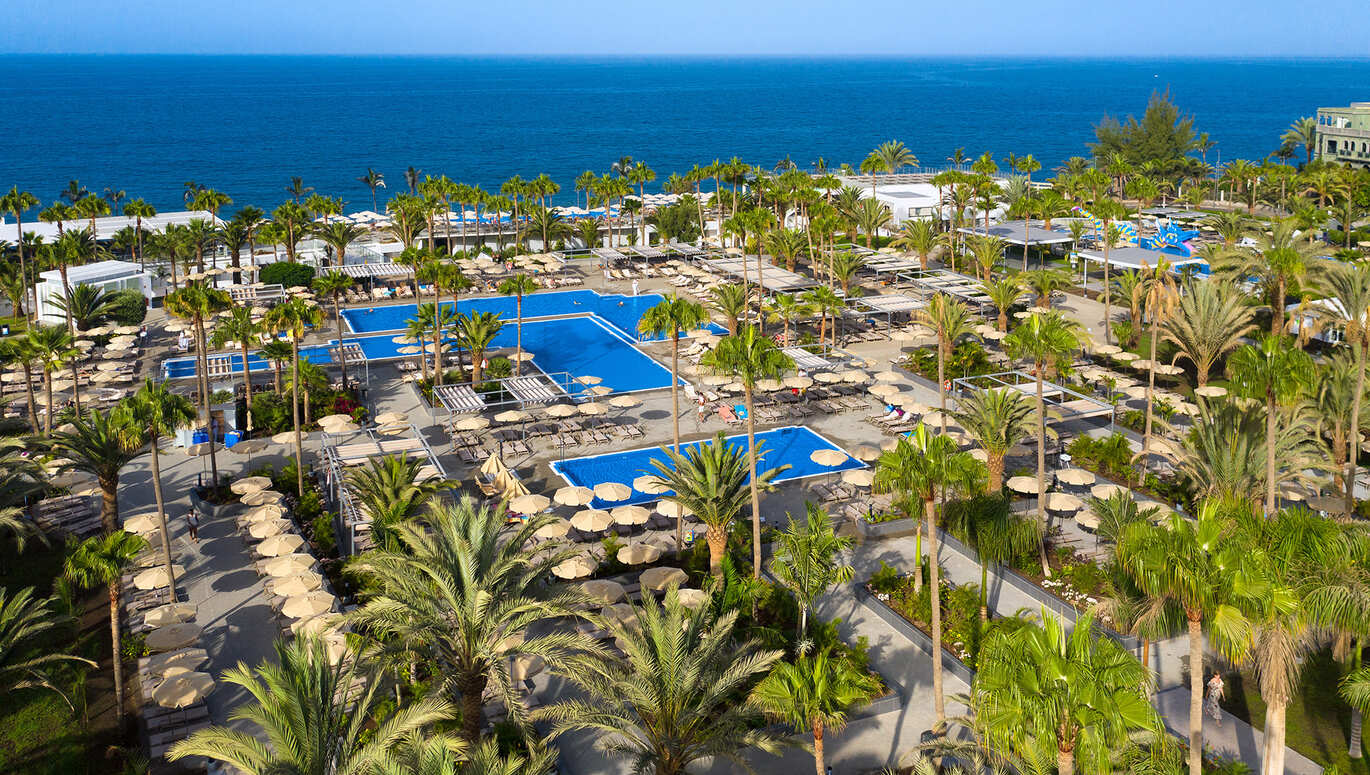 Aerial view of the pool area with the sea behind, surrounded by deck chairs and umbrellas with many palm trees dotted around on a clear, sunny day.
