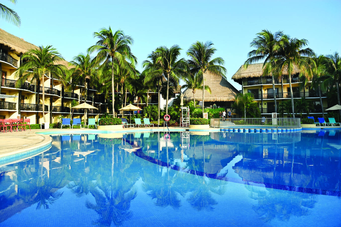 View of the pool with sun loungers and umbrellas around the pool side with many palm trees dotted around and the hotel buildings visible through the trees with straw roofs and the blue sky above.