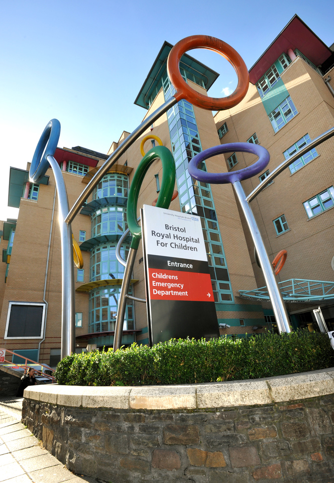 Bristol Royal Hospital For Children, black, white and red sign within a colourful sculpture known as 'Lollypop-Be-bop on a low wall structure with a low hedge and the hospital building behind.