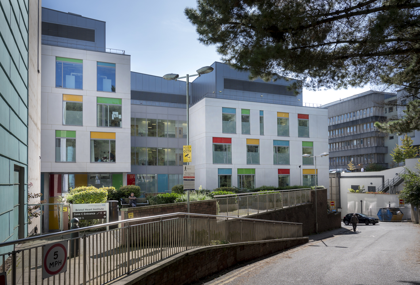 "Bristol Heart Institute. A 4 storey modern, white building with lots of windows and a coloured section on each. There is a small patio area at the front of the building with a bench and decorative bushes. There is an NHS sign outside the building which reads 'Bristol Heart Institute'"