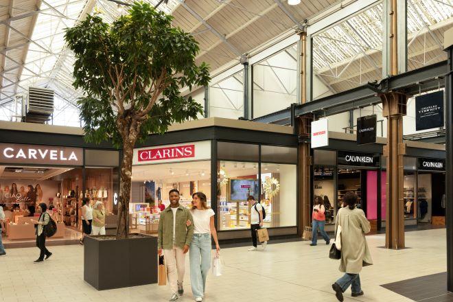 "Interior of Swindon Designer Outlet showing stores including Carvela and Clarins, with a large potted tree in the center and shoppers walking through the bright, spacious area"
