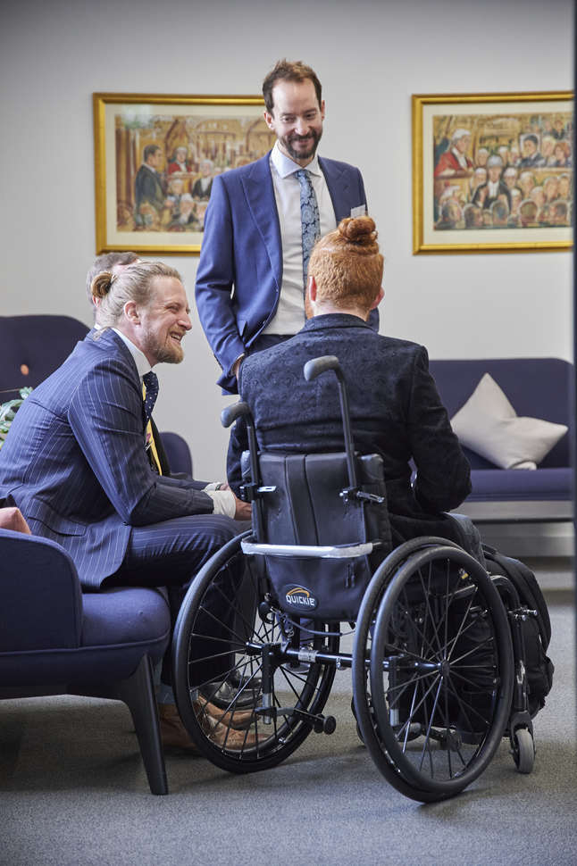Three men in blue suits in a lounge. One is sitting in a wheelchair.