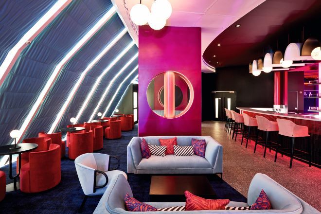 "A lounge bar with a pink theme. In the centre there is a seating area with two sofas and a chair facing inwards towards a small coffee table. To the left there are a row of 4 circular tables with 4 chairs around each. There is a circular lamp on each table. To the right there is a bar with 6 stools."