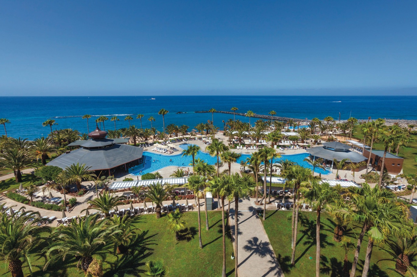 Aerial view of the resort, you can see the two pools with snack/drinks huts with sun loungers and umbrellas around, surrounded by many palm trees, greenery and the sea beyond. It is a lovely clear, sunny day.