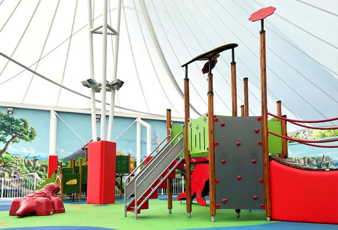 "Indoor children’s play area with colorful climbing structures, slide, and soft flooring under a large white canopy"