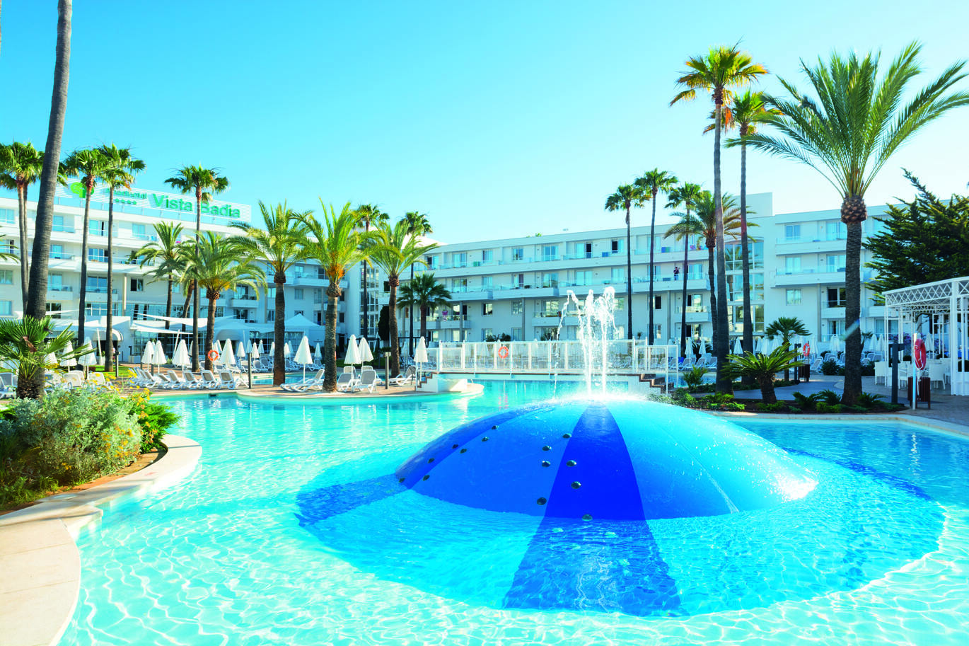 View from the back end of the pool with a fountain in the pool and small climbing handles up the side of the fountain. The pool has sun loungers and umbrellas around with many palm trees dotted around the pool area and the big white hotel building surrounding on a clear sunny day.