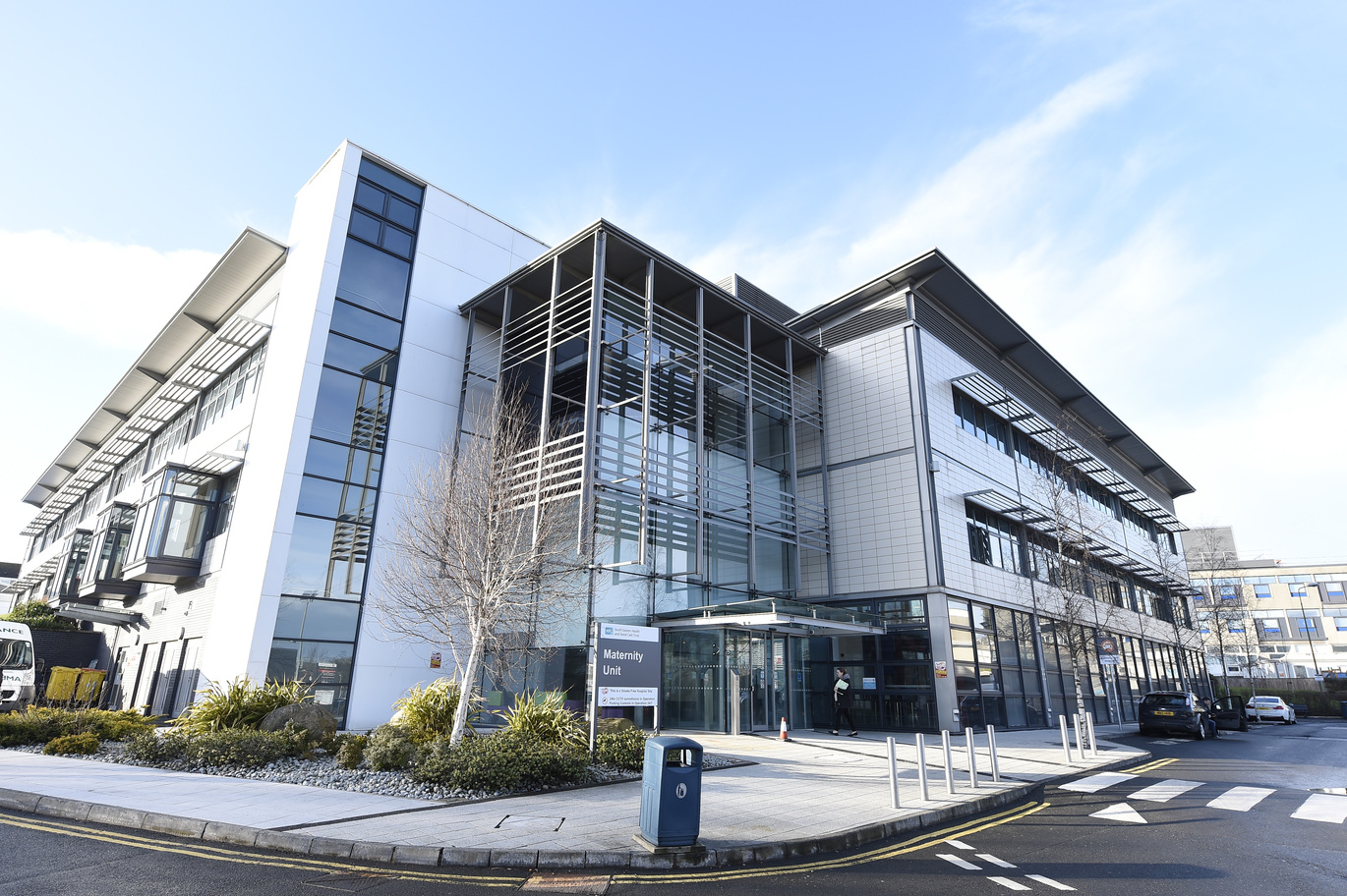 "The Maternity Unit at Ulster Hospital. It is a large, modern building with glass and metal panelling across multiple floors. There is a drop off point at the front of the hospital where a couple of cars are parked. There is a zebra crossing leading to the main entrance and a sign reading 'Maternity Unit'"