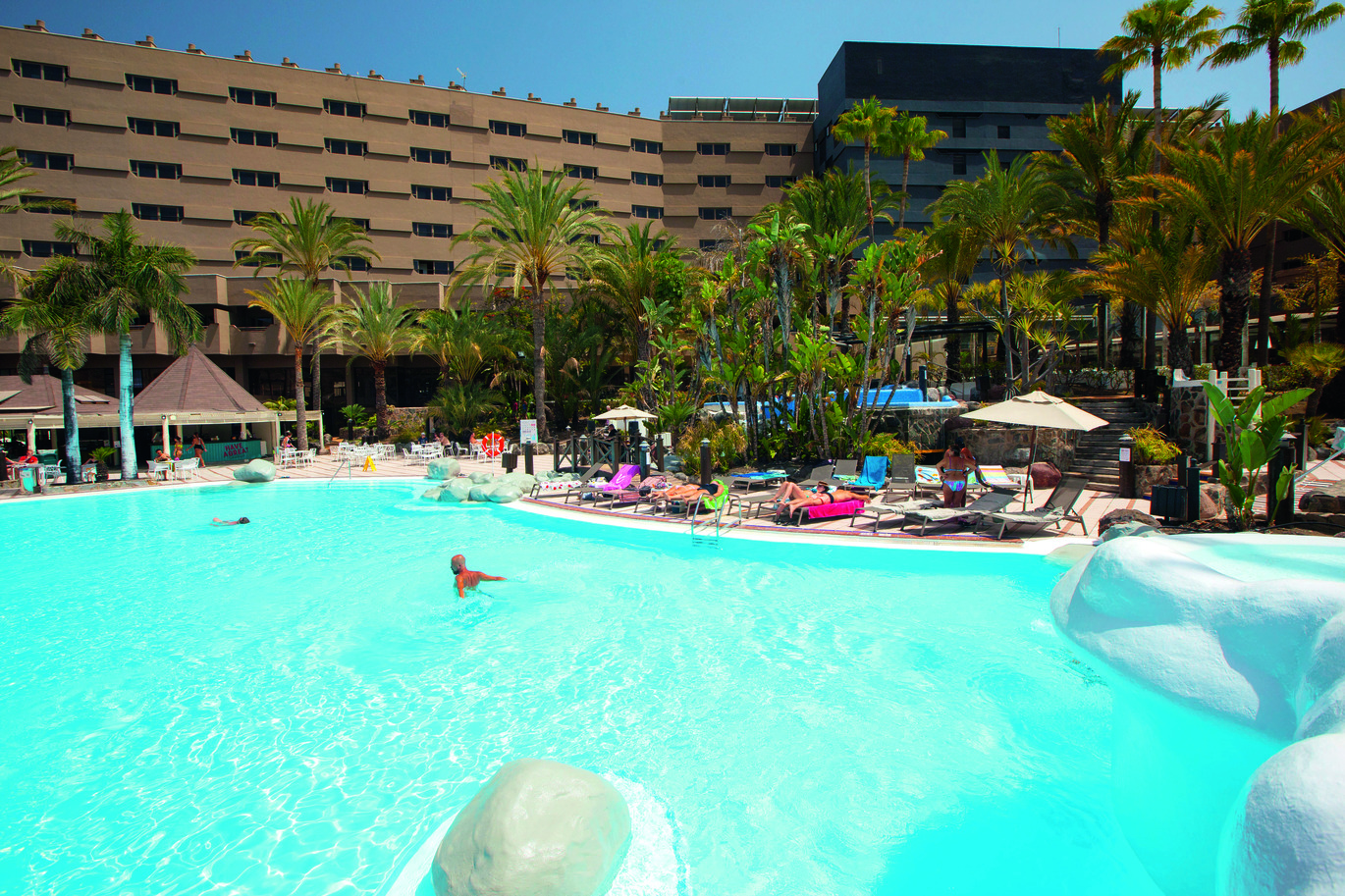 View of the pool area with people in the pool and around on sun loungers, with many palm trees around and the hotel building behind with the blue sky above.
