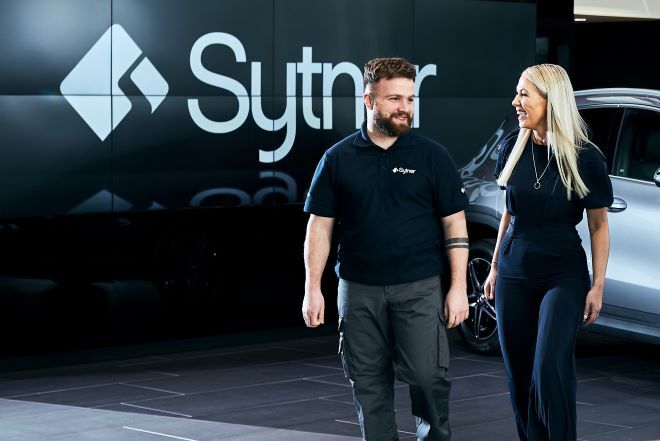"two people stand in front of a silver car, with a large sign that says Sytner in the background"