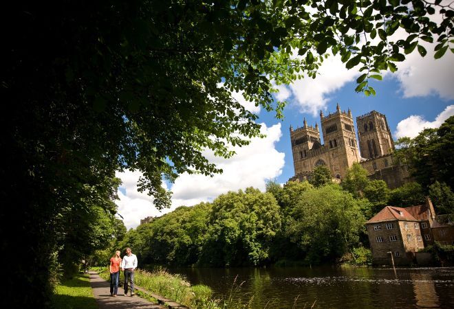 "A male and female walk along a path next to a river, with Durham Cathedral in the background amongst trees"