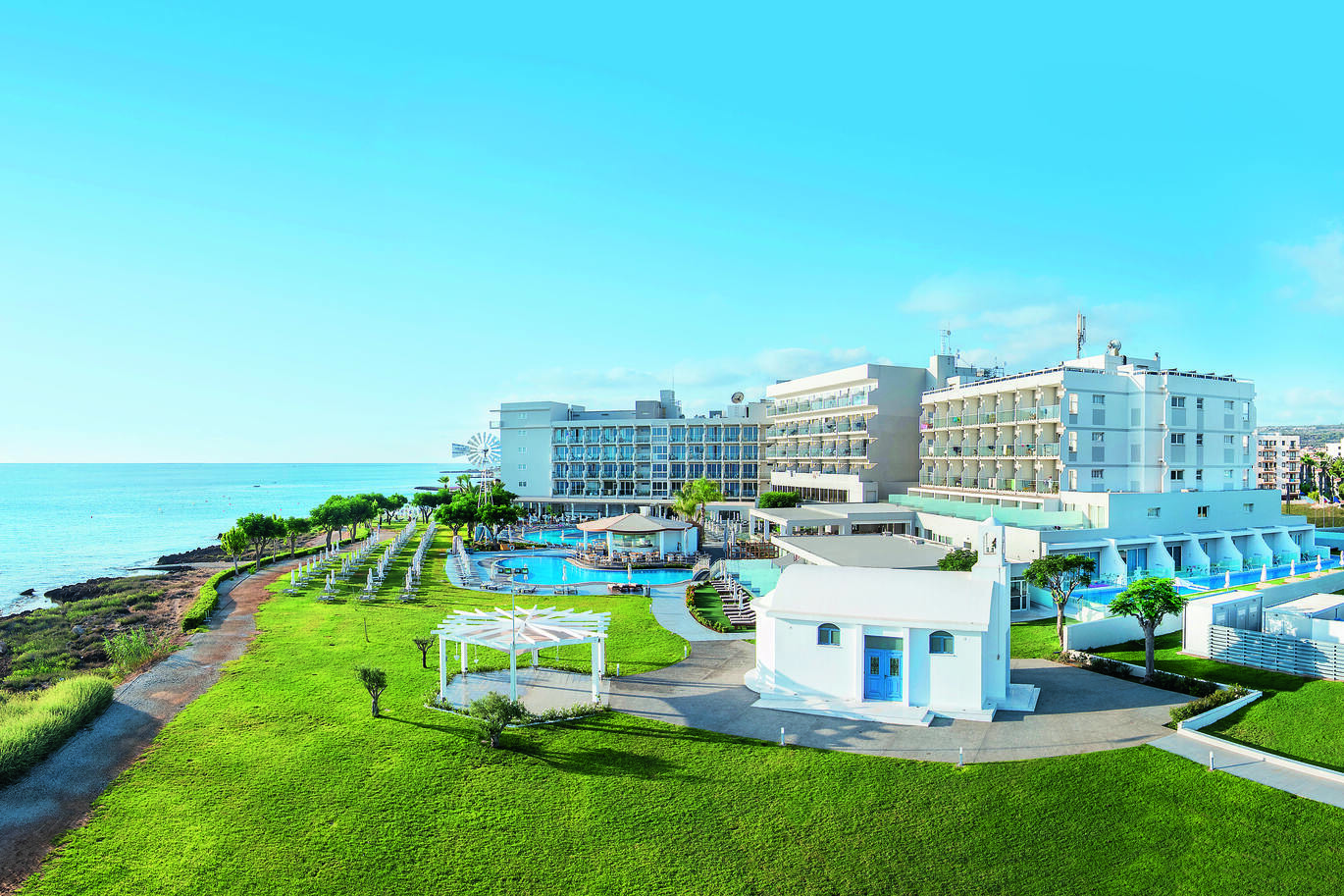 Aerial view of the hotel complex right on the sea front with green lawns all around, the pool in the middle and white hotel buildings surrounding with the blue sky above.