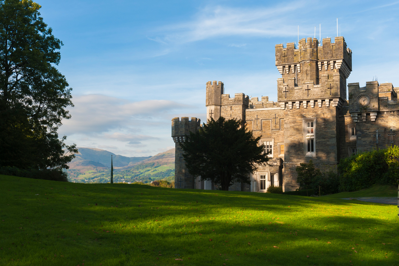 View of the 19th century Wray Castle over the lawn with trees in front and hilly scenery behind.