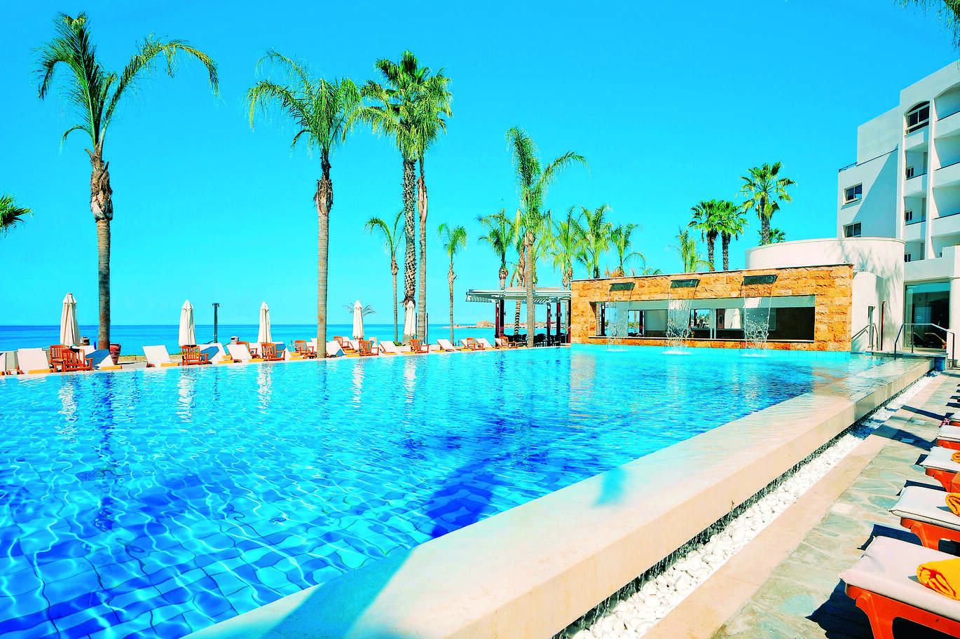 View of the pool overlooking the sea, surrounded by deck chairs and umbrellas on a clear, sunny day.