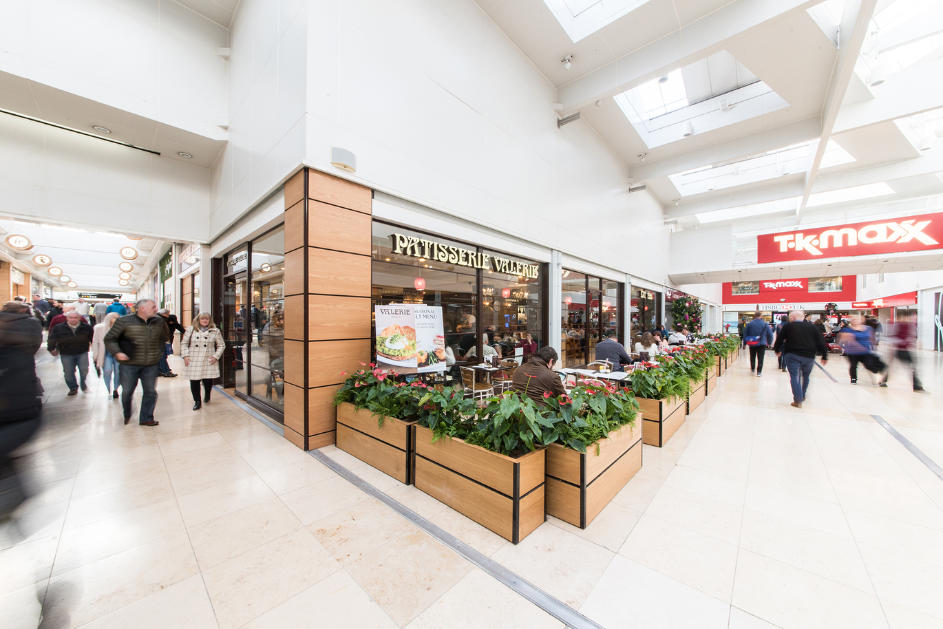 "Patisserie Valerie located inside the shopping centre. There are plants in raised planters outside the restaurant. TK Maxx is located to the right. There are people blurred in the image."