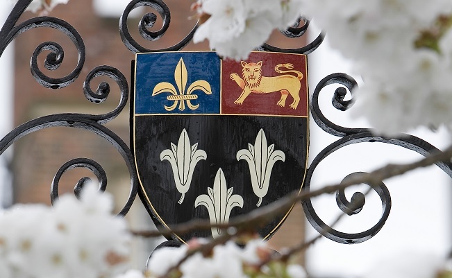 "Photo of the Eton College Coat of Arms. It is displayed on a gate with white blossom branches displayed in front of it."