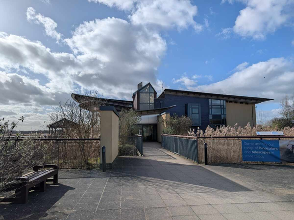 A modern visitor centre building sits at the end of a paved walkway with railings, under a bright sky with scattered clouds