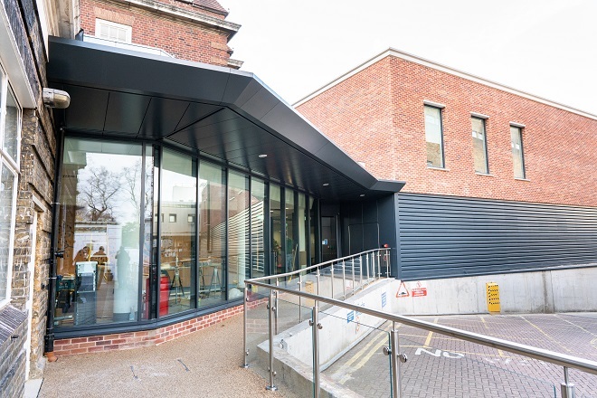 Photo of entrance to Royal Brompton Diagnostic Centre. There is a handrail to the right side going up the ramp. There are clear glass windows as you arrive towards the entrance doors.