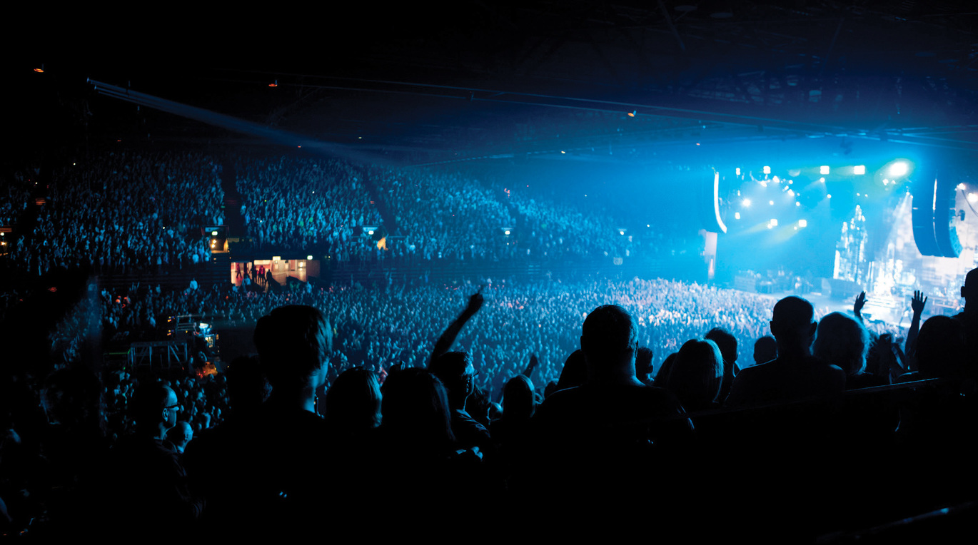 "A photo of inside Genting Arena, the lighting is low around the arena with lights shining out from the stage. The arena is full of people and the photo is taken from the back near the top."