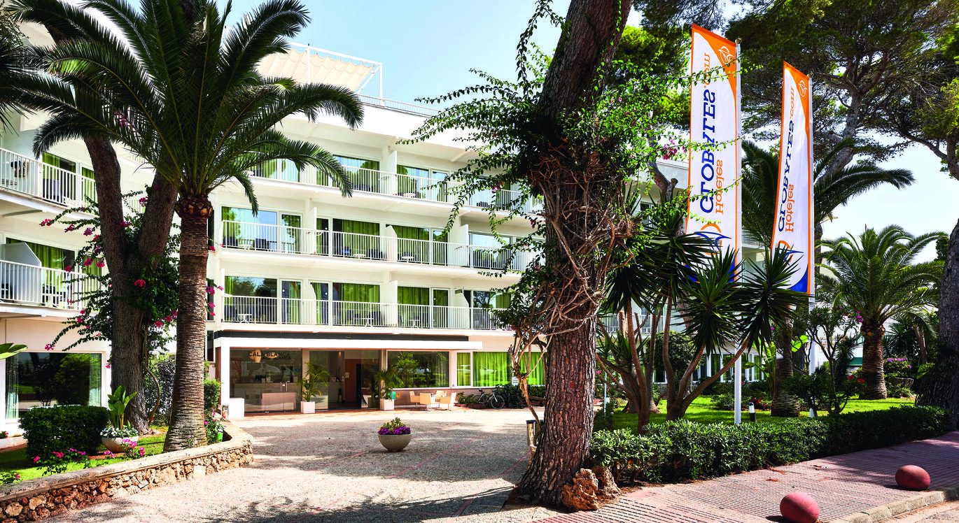 View of the hotel entrance with hotel rooms with balconies above, a gravel courtyard out the front and many trees around.