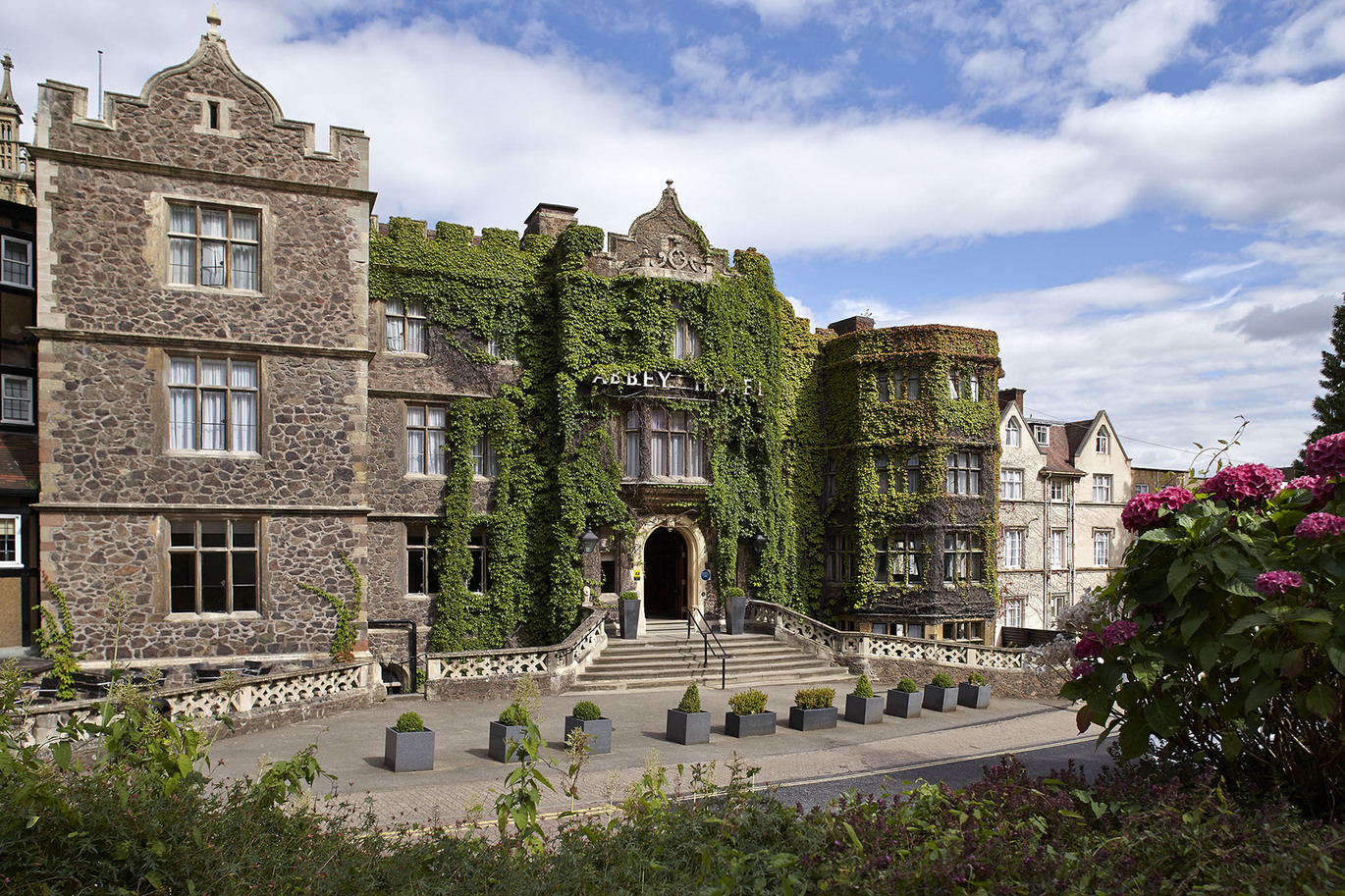 The front of The Abbey hotel, a lovely stone building with the front covered in greenery. Large steps leading up to the front entrance with some large plant pots just below the steps and a wall surrounding the front of the building. Some shrubbery at the bottom of the image with some pink flowers to the right and the lovely blue, slightly cloudy sky above. 