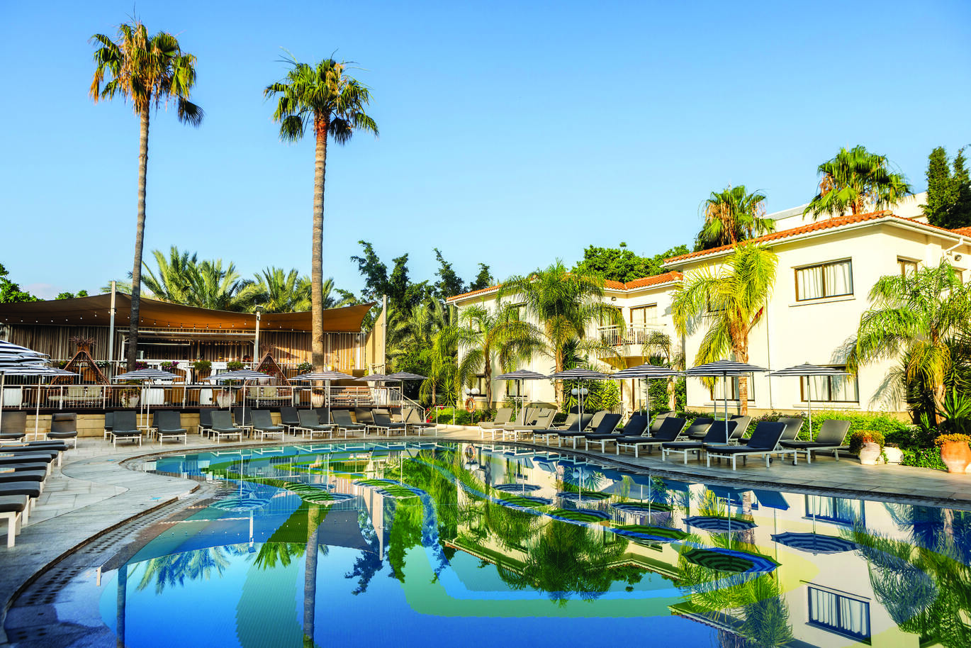 View from the pool area of a number of sun loungers and umbrellas with a small part of some hotel rooms behind on a clear sunny day.