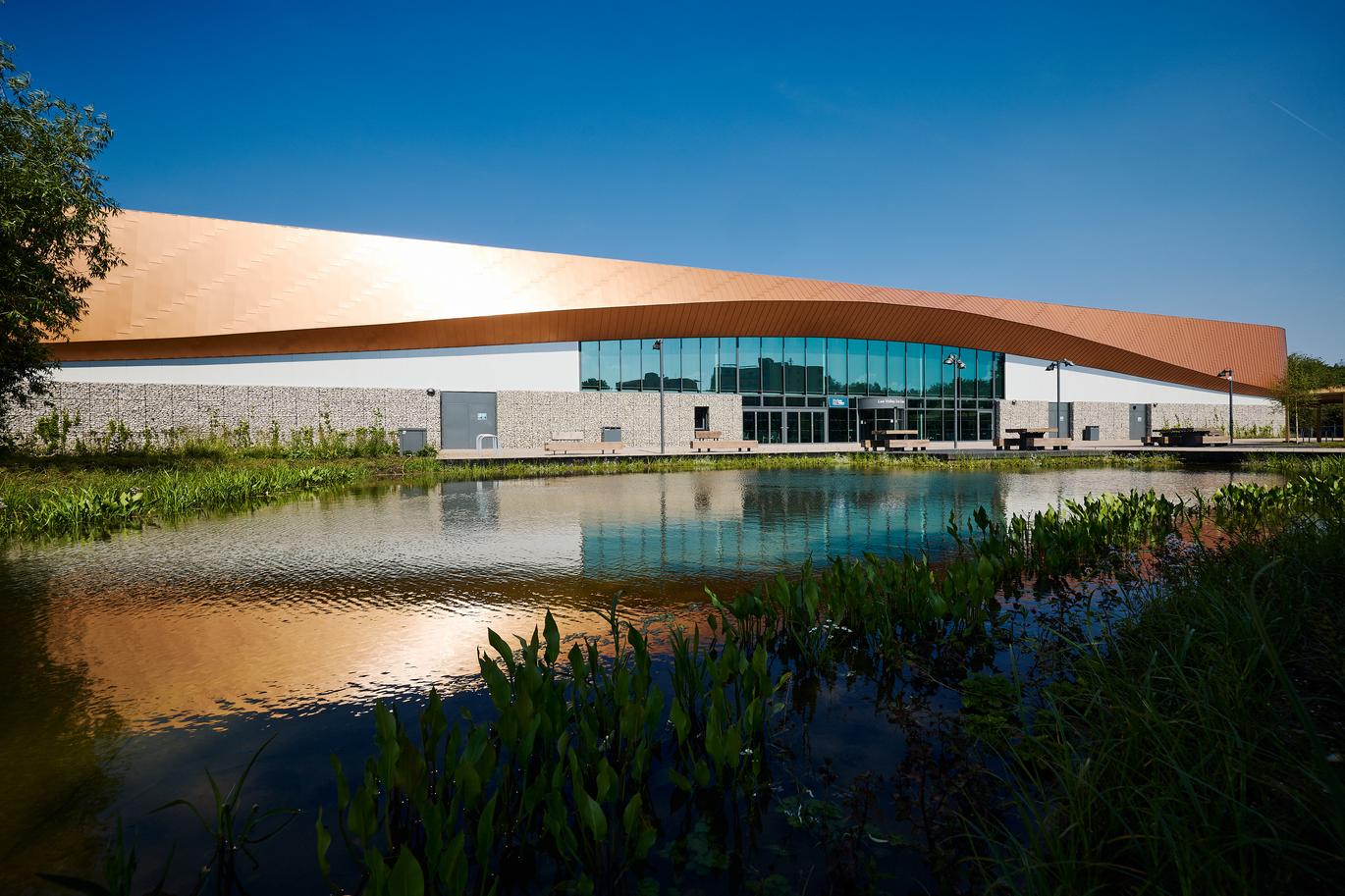 "Modern leisure centre with curved copper-coloured roof and large glass frontage beside a reflective pond, with plants and seating in the foreground."