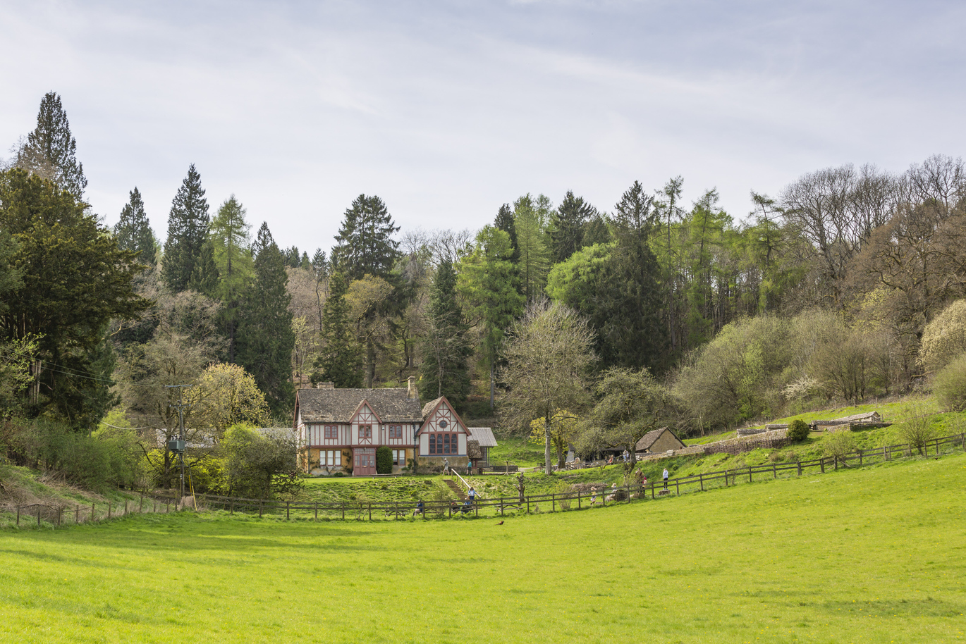 A cottage like house in the middle of a lush green garden with a fence in front separating it from the front field. There are people walking around in the garden and steps on a hill leading up to a higher level of garden and the house. Behind the house are many trees that you cannot see beyond.