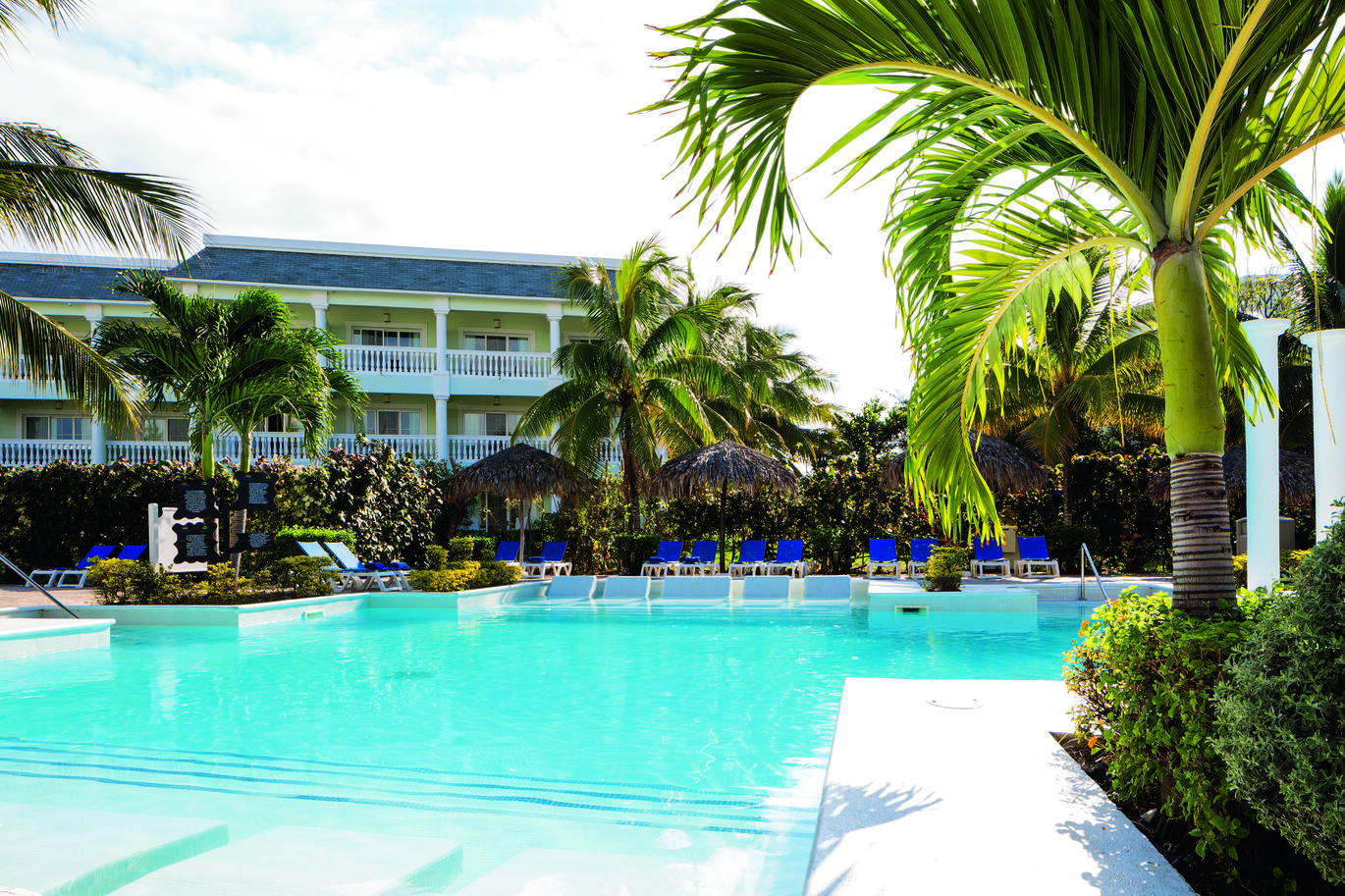 View from the corner of the pool with sun beds and umbrellas surrounding with palm trees dotted around and the hotel building behind.