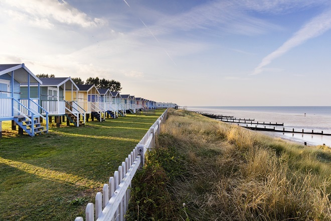 "There are multicoloured beach huts to the left on grass. There is a low white fence between them and the bank down to the sea."