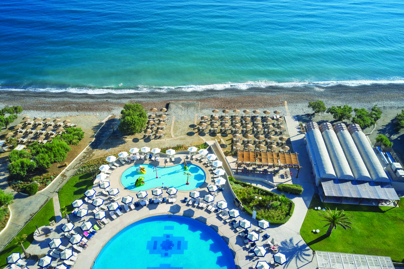 Aerial view of the pool area, surrounded by deck chairs and umbrellas backing on to the stony beach and blue sea.