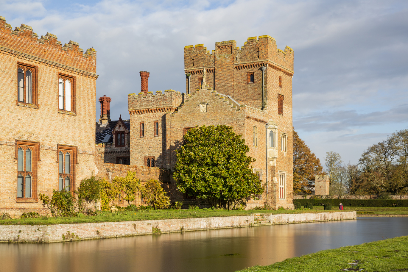 A large, brick, medieval manor house surrounded by a moat with a small piece of lush green lawn and a few trees with a wall dropping down into the moat and some steps within the middle of the wall leading into the moat.