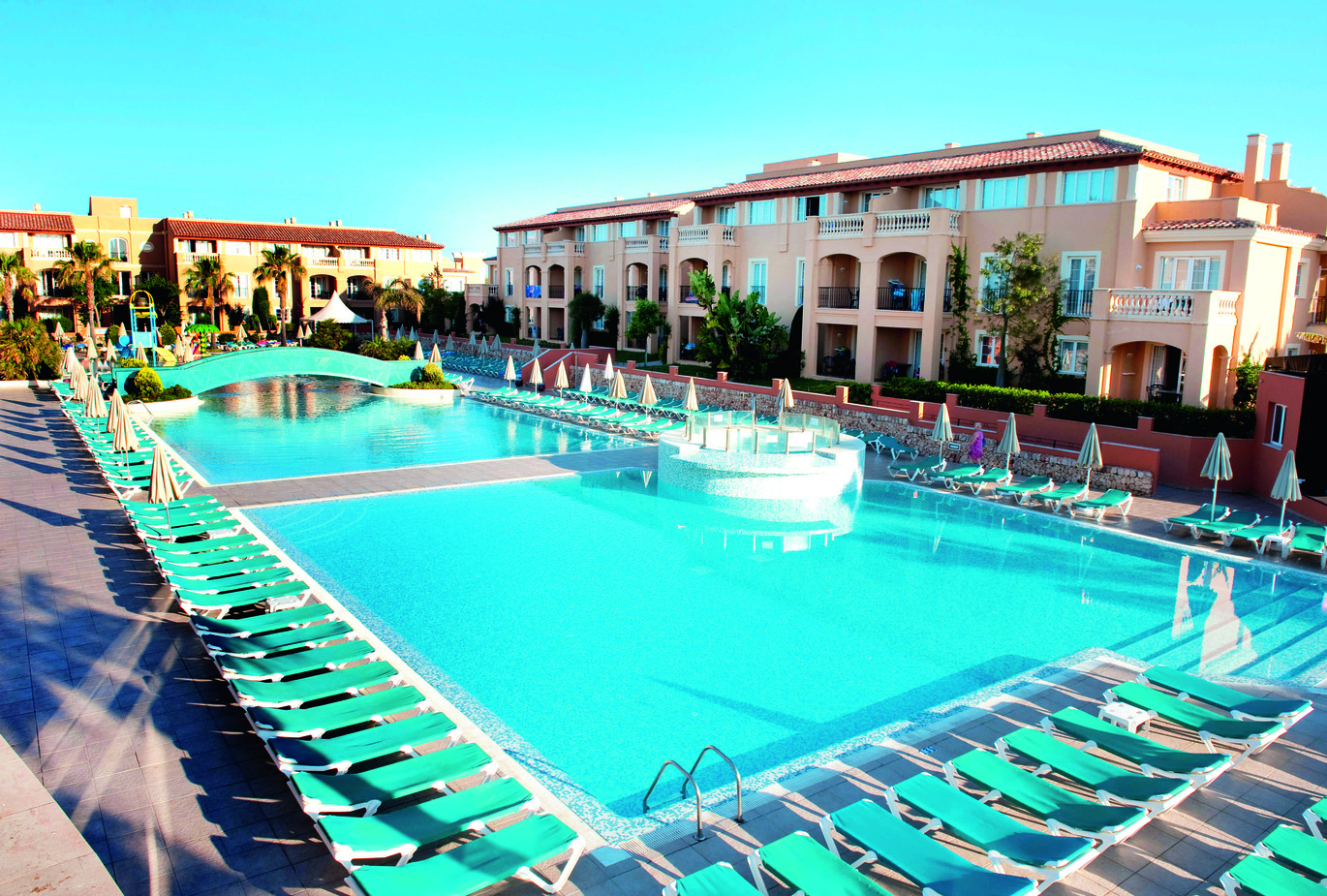 View of the pool area surrounded by many sun loungers, a path through the middle of the pool and a bridge over the pool further up. With clusters of hotel rooms behind and the blue sky above.