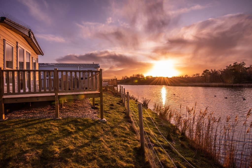 Sunset over a serene lake with a wooden cabin on the left. Warm sunlight reflects on the water, casting a peaceful, golden glow over the scene.