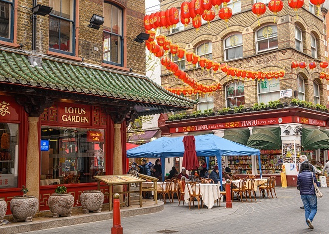 "Lotus Garden is located to the left with plant pots outside and a menu board. There is a blue marquee outside with seating underneath the marquee. There are red lanterns above streamed between buildings."