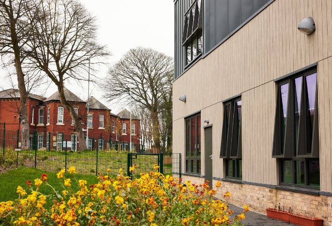 Side view of the building with lawns and flowers outside and brick buildings in the background behind leafless trees.