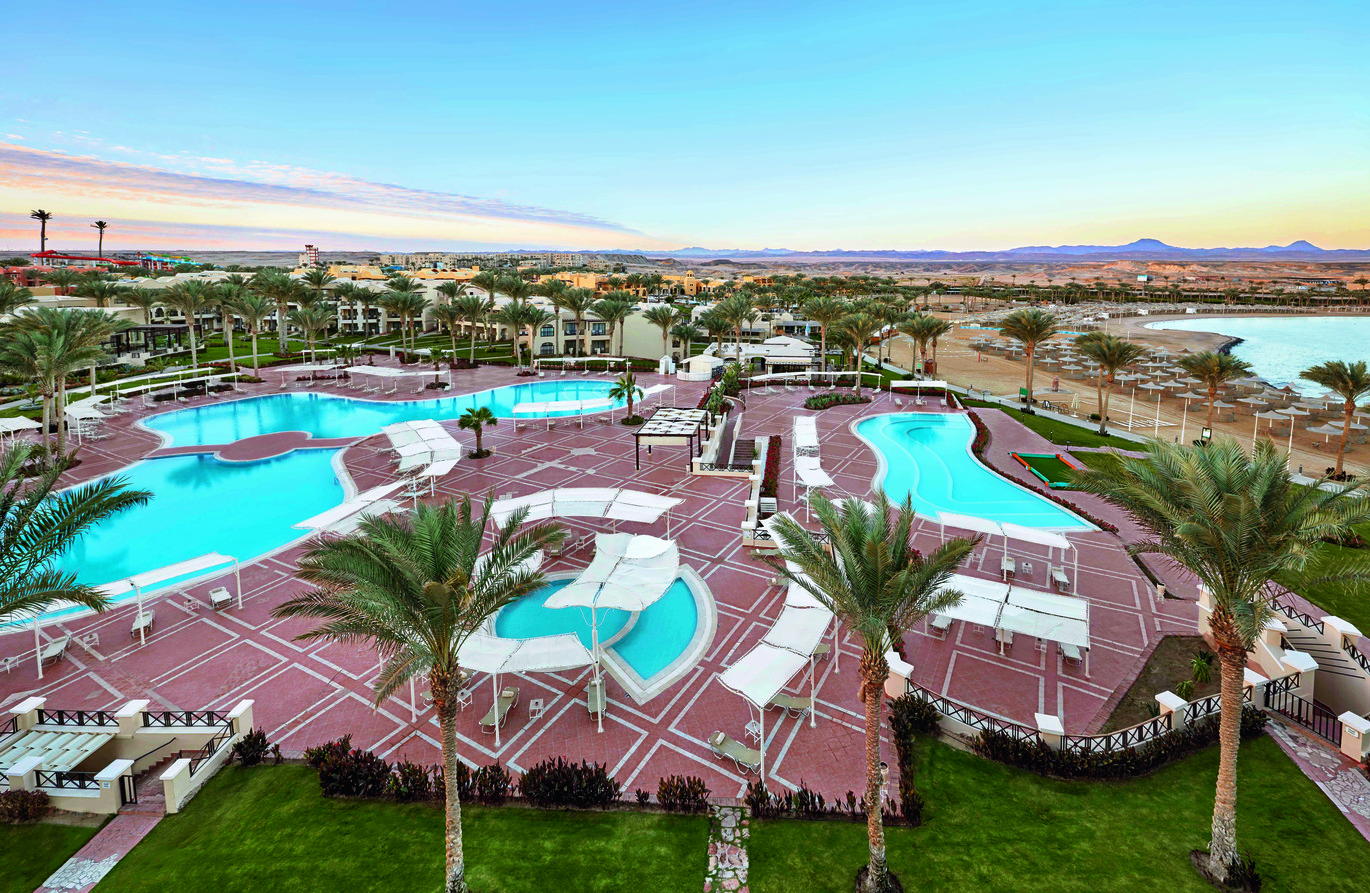Aerial view of the pool area with 3 pools with lawns and palm trees scattered around, the hotel building behind and more buildings beyond. The beach is visible to the side with the sea just in the shot.