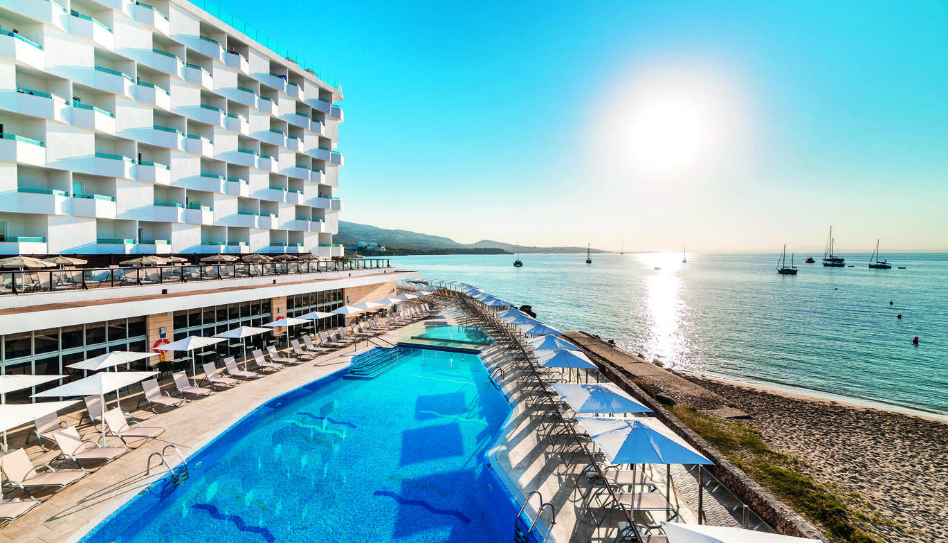 White hotel building with cluster of hotel rooms to the left of picture, a number of sun beds on a higher level below the hotel and then more sun beds on a lower level around the blue pool, all beds have umbrellas above them. A small section of beach to the right of picture and then the sea behind with a number of boats around and the sun shining brightly in the blue sky with some mountains behind.