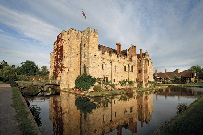"Photo of Hever Castle. There is a moat around the edge of the castle, the castle sits in the middle with trees around the edge. There is a flag at the top of the castle."