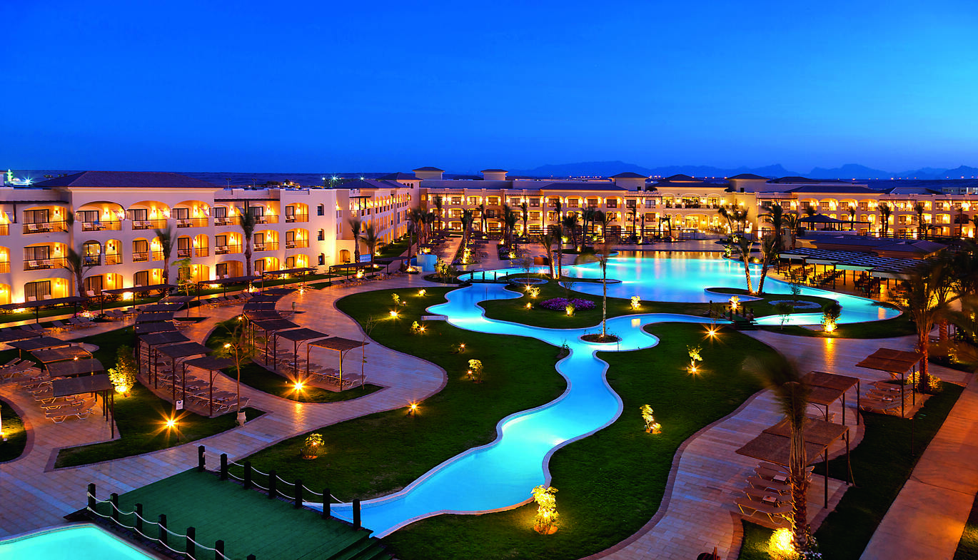 View in the dusk of the large hotel complex with rivers of pools between lawns leading to the main big pool, surrounded by little spot lights with the hotel building surrounding.
