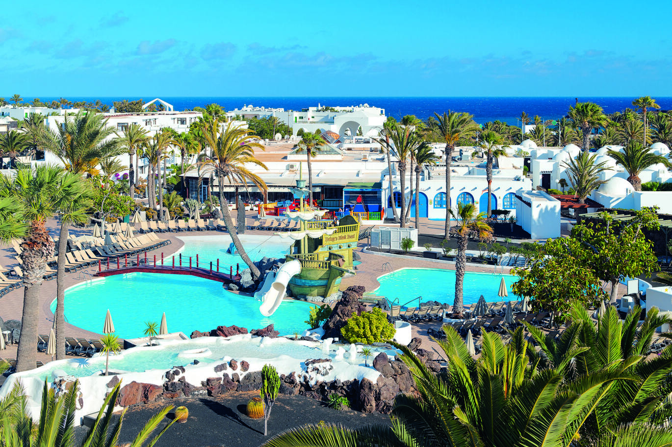 View of the pool area with a pirate ship and slide into the pool on an island in the middle with a bridge over the pool between. There are sun loungers and umbrellas around the pool with palm trees dotted around and the white hotel building behind with the sea just visible in the background.