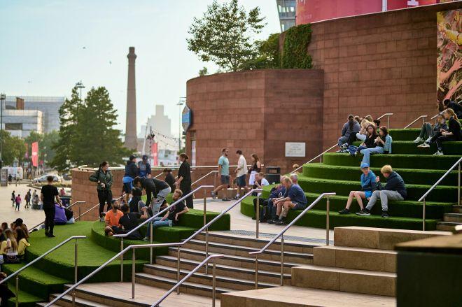 "Tiered seating area at Liverpool ONE with people sitting on grass steps beside a wide pedestrian walkway."